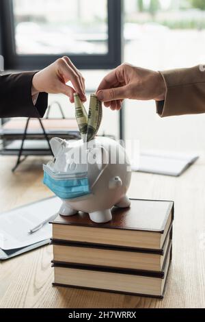 vista ritagliata di avvocato e cliente che mette dollari in piggy banca in maschera medica, anti-corruzione concetto Foto Stock