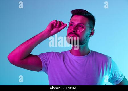 Ritratto corto di uomo bearded che tocca i vetri e guardando attentamente via isolato su sfondo blu in luci al neon Foto Stock