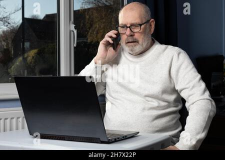 Maschio dai capelli grigi che parla sul telefono cellulare mentre lavora a un computer portatile in un ufficio domestico. Foto Stock