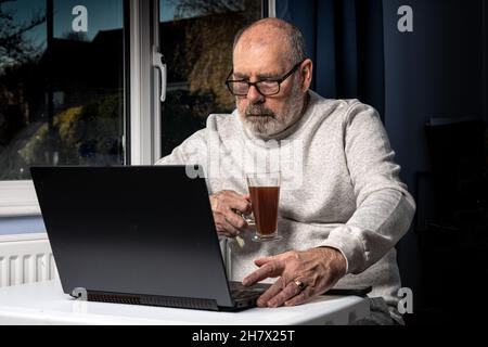 Uomo grigio bearded che beve il caffè mentre lavora su un laptop dal suo ufficio domestico. Foto Stock