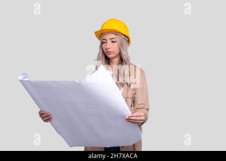 Ragazza costruzione lavoratore Holding House piano in mani guardando esso. Ragazza architetto che tiene Blueprints. Casco rigido giallo. Foto Stock