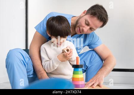 Giovane terapista che aiuta il ragazzo carino che ha paralisi cerebrale, giocando con lo sviluppo giocattolo in clinica di riabilitazione. Foto di alta qualità Foto Stock