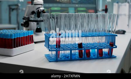 Primo piano delle provette per analisi di laboratorio con sangue sul posto di lavoro. Matracci di vetro riempiti di sostanza rossa o liquido posti su vassoi chimici Vacutainer e microscopio utilizzati in microbiologia Foto Stock