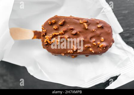 Macro closeup piatto Lay vista dall'alto di una barra gelato al cioccolato alle mandorle su bastone di legno con confezione di plastica bianca sul tavolo da cucina Foto Stock