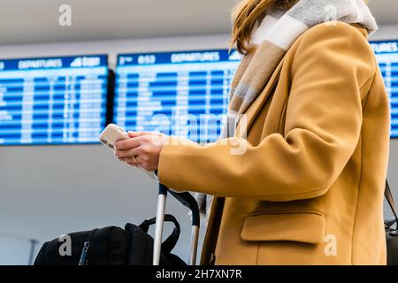 Il viaggiatore donna in elegante cappotto giallo con una sciarpa calda tiene il telefono cellulare e la valigia in piedi contro il bordo di informazioni nel primo piano del terminal dell'aeroporto Foto Stock