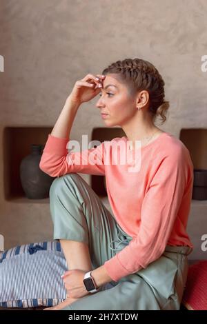 bella donna pensiva in felpa di corallo e pantaloni grigi è seduta in camera Foto Stock