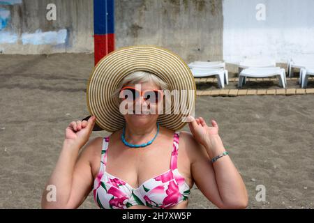 Ritratto di anziani donna sorridente che si trova sulla spiaggia di cappello di paglia e occhiali da sole. Foto Stock