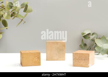 Tre basi quadrate in legno su sfondo bianco e grigio con rami di euclipto con foglie Foto Stock