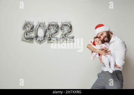 Un padre felice in un velluto rosso Santa cappello sta tenendo la sua figlia del bambino vicino palloncini d'argento nella forma di 2022. Un papà bearded sta abbracciando il suo piccolo Foto Stock