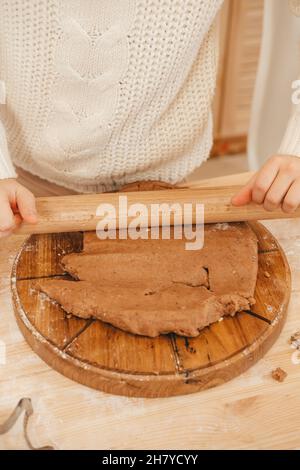 le mani della ragazza tirano fuori l'impasto dello zenzero con un mattarello nella cucina. Ragazza che prepara i biscotti di natale su un tavolo di legno Foto Stock