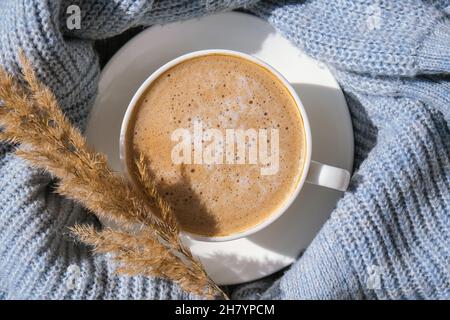 Erba di Pampas e tazza bianca con caffè su maglione blu. Bere cappuccino la mattina della colazione a casa. Disposizione piatta. Sfondo. Estetica. Autunno Foto Stock