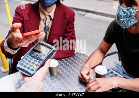 giovane uomo adulto che paga con il telefono cellulare utilizzando un dataphone e la mascherina protettiva waring - nuovo normale Foto Stock