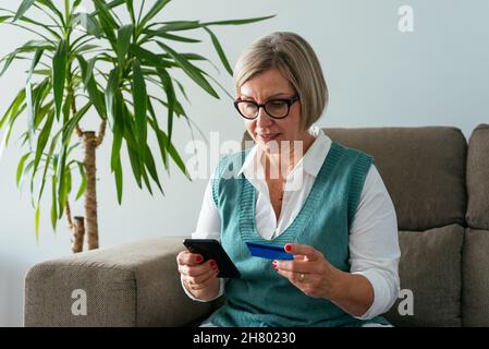 Donna di mezza età seduta sul divano e pagamento per acquisti online con carta di credito e cellulare Foto Stock