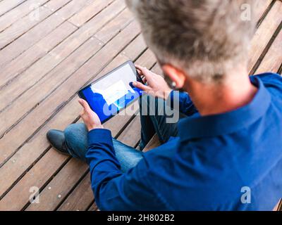 Alto angolo di raccolto anonimo maschio seduto su panca di legno e controllare le notifiche sul tablet Foto Stock