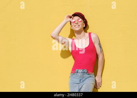 Giovane modello non binario sorridente mentre si posa all'aperto appoggiandosi contro una parete gialla. Foto Stock