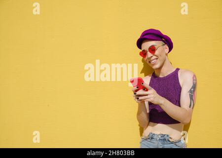 Felice persona non binaria sorridente mentre si utilizza un telefono cellulare all'aperto contro un muro giallo. Foto Stock