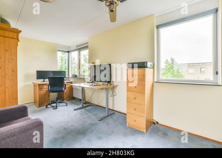 Posto di lavoro con moderno computer sul tavolo in sala luminosa con comoda poltrona e libreria in legno con letteratura vicino alla finestra in appartamento Foto Stock