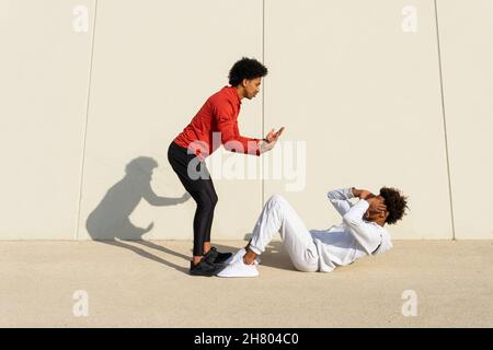 Vista laterale di un istruttore afroamericano che aiuta l'uomo etnico a fare scricchioli sul marciapiede vicino al muro bianco sulla strada Foto Stock