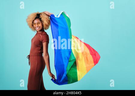 Felice donna afro che detiene una bandiera LGBT orgoglio mentre in piedi su uno sfondo isolato. Foto Stock