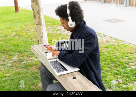 Pensivo studente maschile afroamericano in outerwear e cuffie seduta su panca e prendere appunti in blocco appunti mentre fare i compiti su netbook in parco Foto Stock