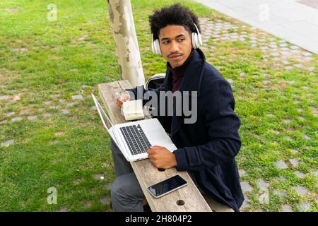 Pensivo studente maschile afroamericano in outerwear e cuffie seduta su panca e prendere appunti in blocco appunti mentre fare i compiti su netbook in parco Foto Stock