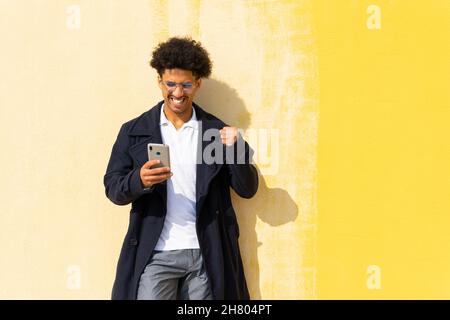 Felice africano americano maschio che alza il pugno mentre legge le buone notizie sul cellulare mentre si alza vicino al muro giallo Foto Stock