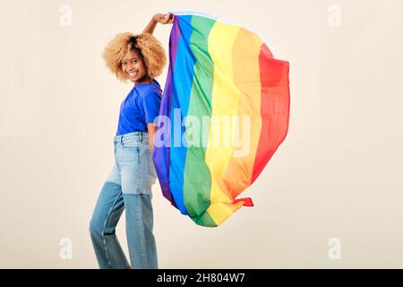 Donna afrofroica sorridendo mentre tiene la bandiera lgbtq di orgoglio gay su uno sfondo isolato. Foto Stock