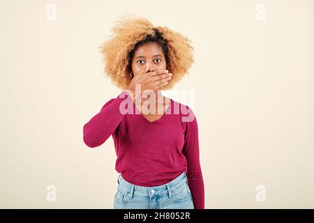 Giovane donna con acconciatura afro che guarda scioccato mentre coprendo la bocca con le mani. Foto Stock