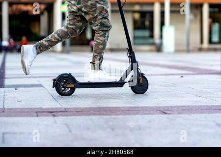 Vista laterale del raccolto irriconoscibile maschio in trendy pantaloni militari e sneakers che rotolano scooter elettrico mentre si cammina sulla strada della città Foto Stock