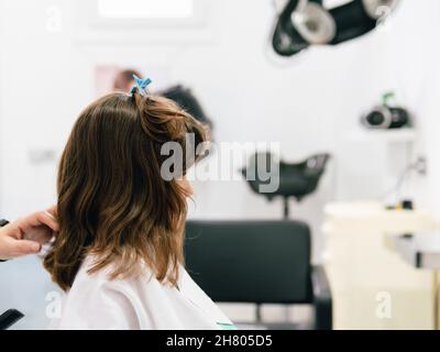 Crop parrucchiere toccare i capelli della ragazza avvolta in cape durante la procedura di acconciatura nel salone professionale Foto Stock