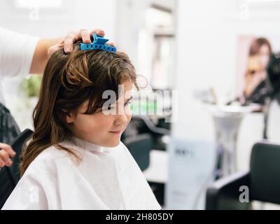 Crop parrucchiere toccare i capelli della ragazza avvolta in cape durante la procedura di acconciatura nel salone professionale Foto Stock