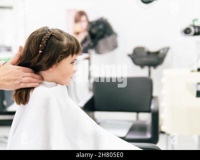 Crop parrucchiere toccare i capelli della ragazza avvolta in cape durante la procedura di acconciatura nel salone professionale Foto Stock