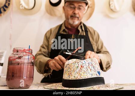 Ritagliare talentuoso artista maschile maturo in abiti casual e grembiule utilizzando pennelli, mentre dipingere su elegante cappello di paglia durante il lavoro in studio creativo Foto Stock