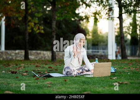 Corpo pieno di ragazza araba allegra in hijab ondeggiare mano mentre si ha video chat via laptop sul prato erboso nel parco Foto Stock