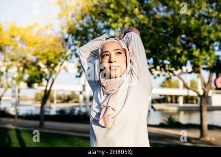 Piacevole femmina araba in hijab stretching triceps mentre si riscalda prima di allenamento fitness nel parco Foto Stock