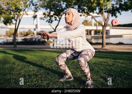 Corpo completo di determinata femmina musulmana in hijab facendo squat allungando le braccia in avanti per l'equilibrio durante l'allenamento all'aperto Foto Stock