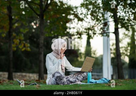 Corpo pieno di ragazza araba allegra in hijab ondeggiare mano mentre si ha video chat via laptop sul prato erboso nel parco Foto Stock