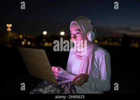 Positiva femmina araba in hijab utilizzando cuffie wireless e guardare video su netbook con luce viola nel buio Foto Stock