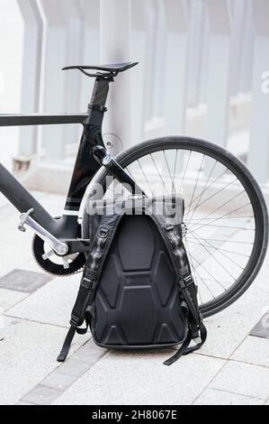Zaino nero posto vicino ad una parte della bicicletta moderna parcheggiata in strada in città Foto Stock