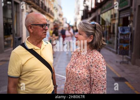 Coppia matura di turisti in abbigliamento casual che si guardano l'un l'altro mentre si trova su un sentiero lastricato tra gli edifici in città Foto Stock