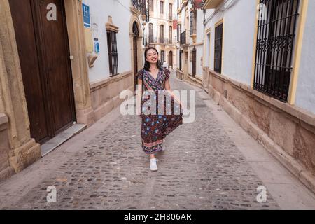 Pieno corpo di allegro viaggiatore asiatico femminile in abito guardando la macchina fotografica mentre si trova in piedi su strada lastricata vicino vecchie case Foto Stock