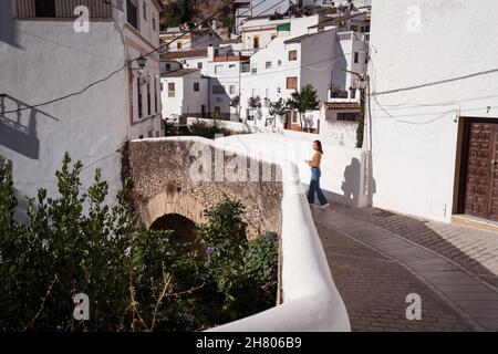 Vista laterale del viaggiatore anonimo lontano passeggiando sul sentiero lungo recinzione vicino a vecchie case residenziali nella soleggiata città antica Foto Stock