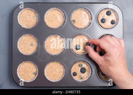 Vista dall'alto della persona anonima del raccolto che mette i mirtilli sopra i muffin crudi nella teglia da forno Foto Stock