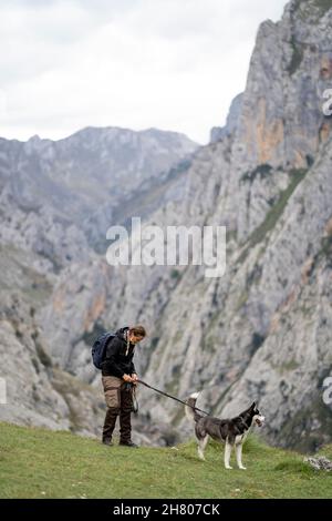 Sorridente signora in abiti caldi e zaino camminare con obbediente Husky su erba verde, mentre si cammina nella valle montuosa Foto Stock