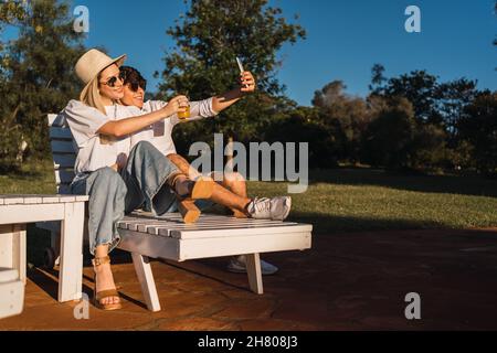 Amando maschio e femmina in occhiali seduti sul lettino vicino prato verde e prendendo auto ritratto mentre godendo l'estate Foto Stock