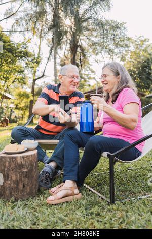 Corpo pieno di uomo anziano positivo in abiti casual e occhiali che mostrano foto sullo smartphone a sorridente moglie mentre si riposa sui lettini nel parco verde Foto Stock