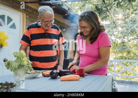 Donna anziana positiva in abiti casual sorridendo e tagliando verdure fresche mature mentre preparano insalata sana con marito sulla terrazza di casa in c Foto Stock