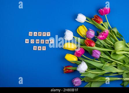 Tessera giorno Happy womens. Lettere in legno. Mix di tulipani primaverili fiori. Sfondo blu con fiori tulipani primo piano diversi colori. Fiore di primavera multicolore. Regalo. Rosso, rosa, bianco e giallo. Foto Stock