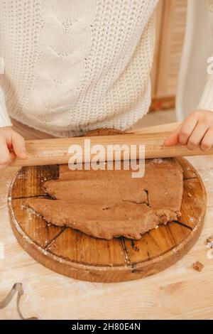 le mani della ragazza tirano fuori l'impasto dello zenzero con un mattarello nella cucina. Ragazza che prepara i biscotti di natale su un tavolo di legno Foto Stock