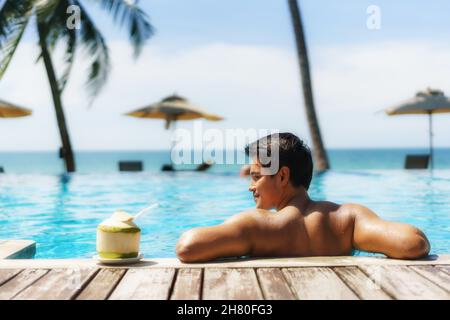 Concetto di vacanza di viaggio d'estate, viaggiatore asiatico uomo con cocco relax nel lussuoso resort a sfioro piscina hotel con spiaggia di mare e palme sfondo Foto Stock